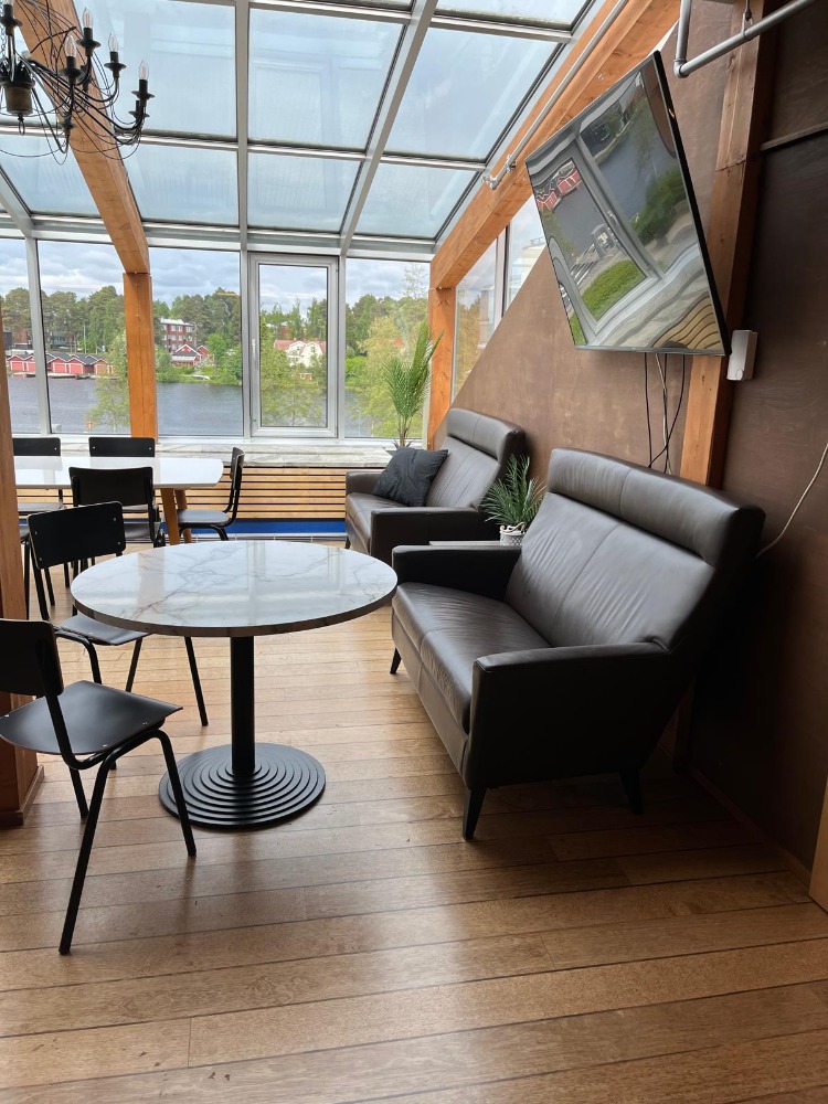 Koskikokous-talvipuutarhan lounge-alue: harmaa nahkasohva, marmoripintainen pyöreä pöytä ja näkymä joelle